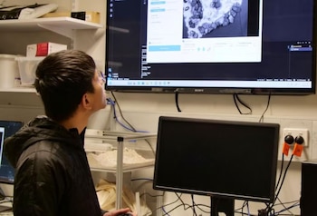 Un hombre con gafas observa una pantalla grande con imágenes microscópicas y código, junto a un monitor de escritorio y cables