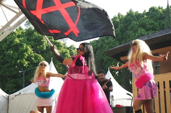 Tres personas disfrazadas de rosa actúan. Una ondea bandera negra con "A" roja. Dos rubias visten de rosa. Hombre toca instrumento. Fondo con árboles y carpas blancas