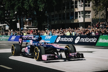 Un monoplaza de carreras azul y rosa con un piloto a bordo transita por una calle urbana, rodeado de espectadores y vallas con logotipos de patrocinadores