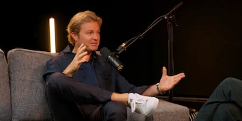 Nico Rosberg, hombre rubio, sentado en un sofá gris, gesticula con las manos. Viste camisa azul oscuro y zapatillas blancas. Un micrófono está frente a él y una luz naranja vertical en el fondo oscuro