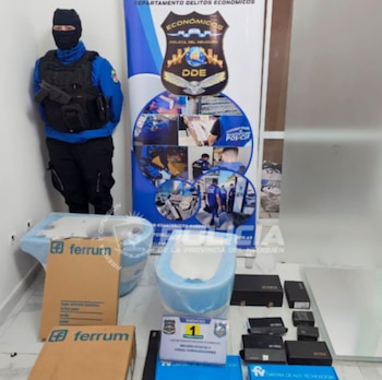 Oficial de policía con pasamontañas, chaleco táctico y arma. Junto a un banner policial, en el suelo hay dos sanitarios, cajas de cartón y pequeños dispositivos electrónicos