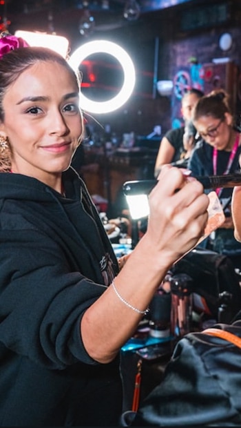 Karina Puente, con una sudadera negra y una pulsera, sonríe a la cámara mientras aplica maquillaje con una brocha, con una luz anular brillante detrás