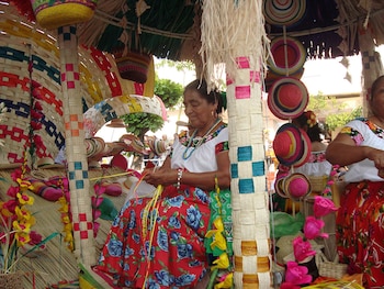 Dos mujeres con vestimenta tradicional, una elabora artesanías con tiras de palma, rodeadas de sombreros, canastas y estructuras de palma tejida multicolores