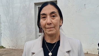 Primer plano de una mujer de cabello oscuro, vestida con chaqueta clara sobre camisa negra y collar, con una pared blanca y una puerta al fondo