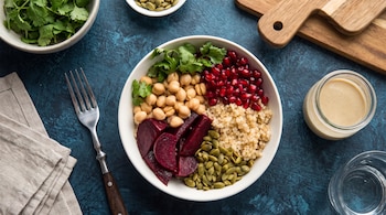 Bowl blanco con quinoa, garbanzos, remolacha, granada, semillas de calabaza, cilantro, tenedor, servilleta, aderezo y vaso de agua.