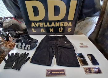 Una mesa blanca exhibe guantes negros, shorts negros, zapatillas Nike, dos teléfonos móviles y un reloj pequeño frente a un cartel de la DDI Avellaneda-Lanús y una motocicleta