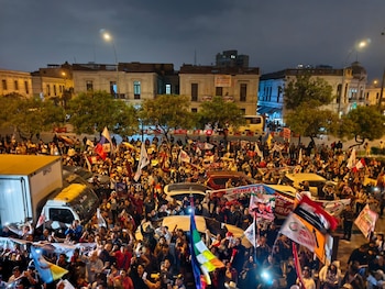 Gran multitud de personas reunidas de noche en una calle urbana con banderas peruanas, pancartas políticas, teléfonos con luces, vehículos y edificios antiguos