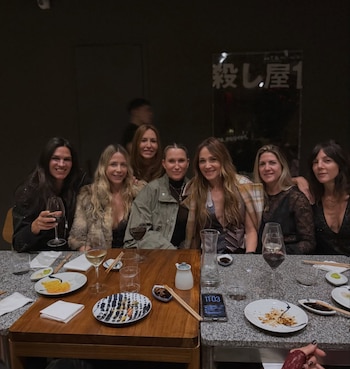 Siete mujeres sonríen sentadas alrededor de una mesa de restaurante, con platos de comida, copas de vino y bebida en el centro