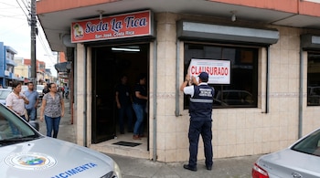 Un funcionario del Ministerio de Salud de Costa Rica, de espaldas, coloca un letrero de "CLAUSURADO" en la ventana de una soda en una calle urbana.