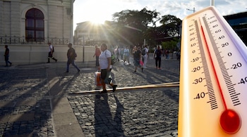 Calle adoquinada, personas caminando, edificios, árboles, sol fuerte y sombras largas. Un termómetro de mercurio muestra grados Celsius y Fahrenheit.