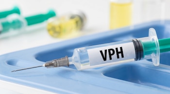 Close-up de una jeringa transparente con émbolo verde, etiquetada como VPH, apoyada en una bandeja médica azul, con viales borrosos al fondo.