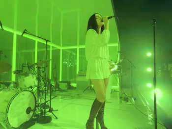 Mujer con vestido blanco y botas oscuras canta en un micrófono en un escenario iluminado con luz verde brillante. Una batería es visible a la izquierda