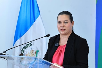 Mujer con blazer negro y blusa rosa habla en un podio con un micrófono y la bandera de Guatemala de fondo