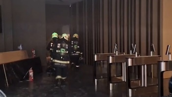 Tres bomberos con cascos, equipos de oxígeno y uniformes oscuros avanzan en un edificio con torniquetes de acceso, un mostrador y un extintor rojo