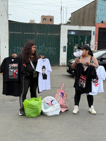 Dos mujeres sostienen ganchos con camisetas del grupo BTS junto a bolsas con ropa colocadas sobre el suelo