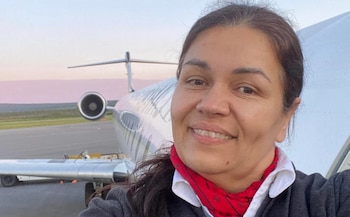 Primer plano de una mujer sonriendo con pelo oscuro, camisa blanca y pañuelo rojo, delante del motor y ala de un avión en la pista al atardecer