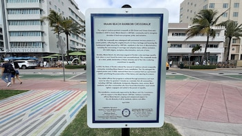 Un cartel blanco en primer plano con información sobre el "Miami Beach Rainbow Crosswalk". Debajo, un paseo peatonal con franjas de colores pastel. Al fondo, edificios, palmeras, una calle y personas