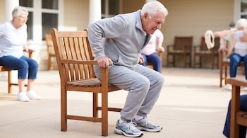 Hombre adulto mayor con ropa deportiva se levanta de una silla de madera. Otras personas mayores sentadas en sillas al fondo, algunas ejercitándose.