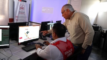 Un hombre con chaleco rojo señala la pantalla de una computadora mientras otro hombre observa, frente a un cartel de Alerta MINEDU.