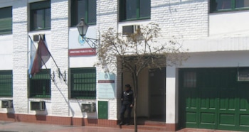 Exterior de un edificio blanco con ventanas verdes, puerta de garaje verde, una persona de uniforme, un árbol sin hojas, y un cartel "Ministerio Público Fiscal" junto a una bandera argentina y otra bordó