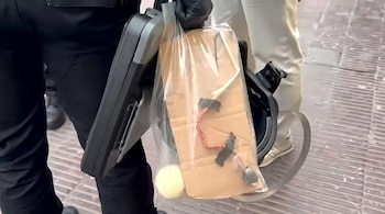 Persona con guantes negros sostiene un maletín negro y una bolsa transparente con un artefacto de cartón, cables y un objeto cilíndrico