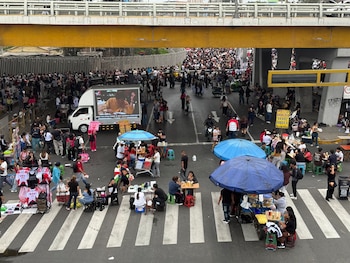 Multitud de personas, puestos de comida y venta de prendas de vestir se ubican bajo un puente vehicular en una calle con paso peatonal