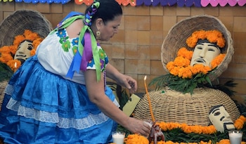 Una mujer con vestido azul y blanco acomoda una veladora encendida junto a canastos con máscaras y flores de cempasúchil