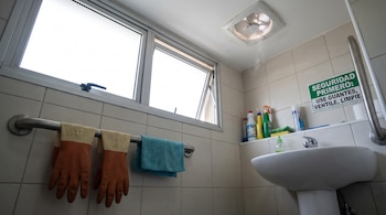 Interior de un baño con baldosas claras, una ventana abierta, un extractor de aire en el techo, un lavamanos, guantes y un paño colgados en una barra y un letrero de seguridad.