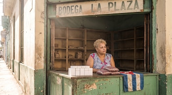 Una mujer atiende tras el mostrador de una bodega con el letrero La Plaza, junto a cajetillas blancas, bolsas con frijoles y la bandera de Cuba.