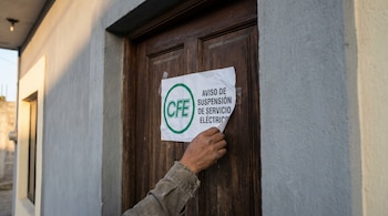 Mano retirando un aviso blanco de suspensión de servicio eléctrico de la CFE de una puerta de madera oscura en una casa mexicana, bajo luz natural de la mañana.