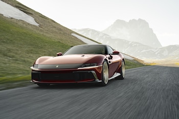 Automóvil deportivo Ferrari color rojo vino en carretera asfaltada con montañas, picos nevados, vegetación y cielo claro de fondo