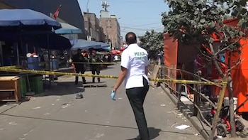 Una agente de Criminalística camina hacia una calle acordonada con cinta de seguridad, frente a puestos comerciales y policías