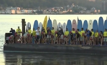 Un grupo de personas en una embarcación oscura, junto a una fila de tablas de surf de colores, sobre un cuerpo de agua con una ciudad al fondo