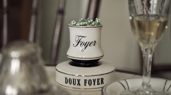 Recipiente cerámico blanco con la palabra Foyer y la frase Doux Foyer, relleno de fósforos con cabezas verdes, junto a una copa con líquido claro.