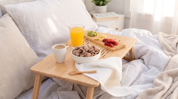 Bandeja de madera en una cama con un tazón de cereales, un vaso de jugo, una taza de café y trozos de fruta.