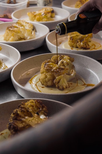 Una mano vierte un líquido oscuro de una botella sobre un trozo de coliflor asada y caramelizada, que reposa sobre una salsa clara en un plato. Varios platos idénticos se ven alrededor