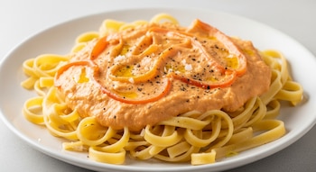 Plato blanco con pasta tagliatelle, salsa anaranjada, rodajas de morrón rojo, pimienta negra, sal en escamas y gotas de aceite de oliva.