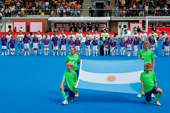 Jugadores de hockey alineados en una cancha azul con niños al frente mientras cuatro jóvenes sostienen la bandera de Argentina