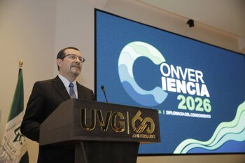 Hombre con traje oscuro y lentes pronuncia un discurso en un podio con micrófono, junto a una pantalla con el texto Convergencia 2026