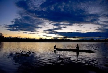 Siluetas de dos personas en una canoa sobre un río al atardecer, con árboles en el horizonte y cielo nublado