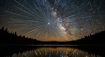 Imagen de larga exposición mostrando una lluvia de meteoros Líridas con la Vía Láctea, reflejada en un lago oscuro, bajo un cielo estrellado y siluetas de árboles.