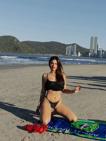 Luana Fernández, con cabello castaño oscuro, arrodillada en la arena, vistiendo un bikini de encaje negro. Detrás, el mar, una montaña verde y edificios modernos