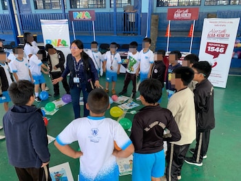 Una mujer con chaleco interactúa con niños en una cancha deportiva; se observan globos y folletos, con banners de PNA y Alerta Alba-Keneth al fondo