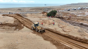 Máquina excavadora amarilla sobre terreno arenoso, sección de carretera interrumpida, personas observando, vehículos estacionados, océano y un muelle en el horizonte