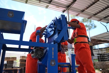 Tres personas con uniformes naranjas y cascos de seguridad operan una máquina industrial azul con un logo institucional. Hay bloques apilados al fondo