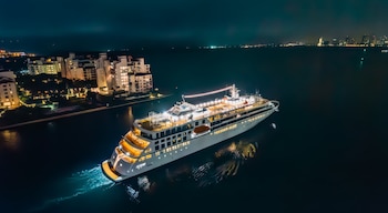 Vista aérea nocturna de un gran barco cruiseferry blanco y amarillo iluminado navegando en aguas oscuras, con edificios costeros y una ciudad en la distancia
