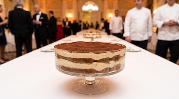 Un tiramisú en un recipiente de vidrio sobre una mesa con mantel blanco, con cocineros y personas vestidas de etiqueta al fondo.