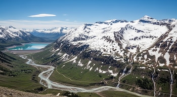 Vista aérea de montañas nevadas y valles verdes con un río serpenteante, una presa y un embalse de agua turquesa bajo un cielo azul con una nube.