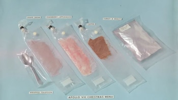 Cuatro paquetes de comida espacial transparentes y una cuchara envuelta se muestran sobre una superficie azul. Las etiquetas identifican bebida de uva, puré, café y pavo
