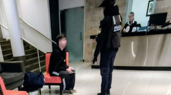 Un hombre sentado en un sillón con el rostro difuminado habla con un oficial de policía en el vestíbulo de un hotel
